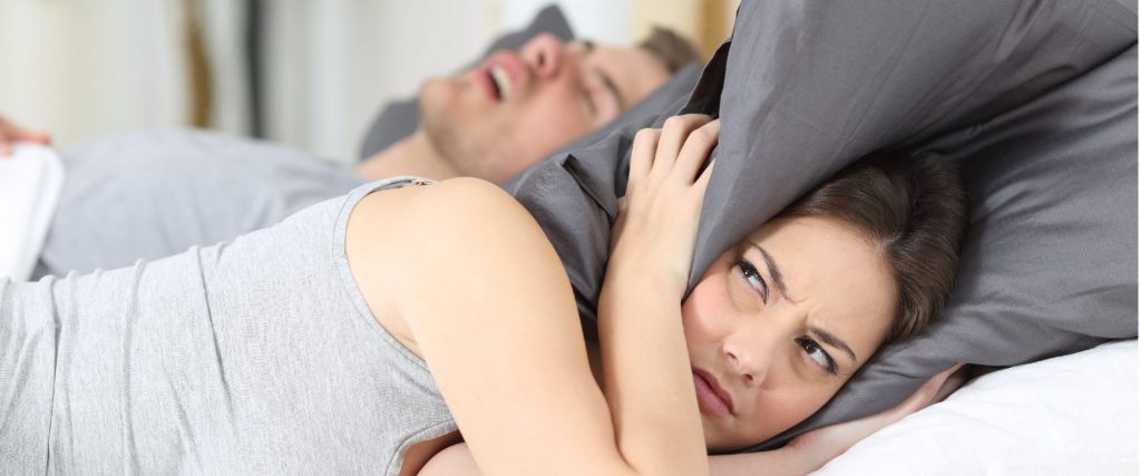 Un ragazzo che dorme russando e la ragazza al suo fianco che si copre le orecchie con il cuscino